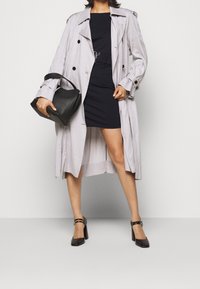 Hellgrauer Trenchcoat mit schwarzen Knöpfen und gebundenem Taillenbereich, getragen über einem enganliegenden schwarzen Kleid. schwarze Lederhandtasche, braune Blockabsatzschuhe.