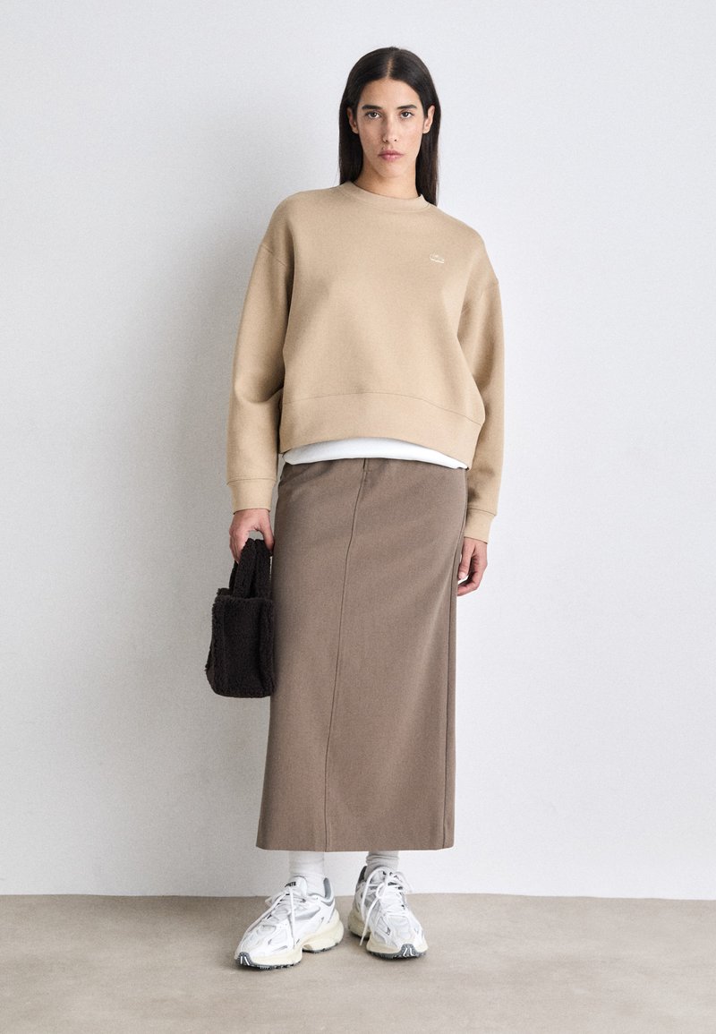 Sweatshirt court marron clair avec des poignets côtelés, associé à une longue jupe marron. Chaussettes blanches et baskets épaisses. Petit sac à main noir.