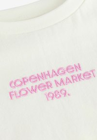 Valge puuvillane T-särk roosa tikandiga tekstiga: "KOPENHAAGENI LILLETURG 1989." Ribaga meeste krae ja pehme tekstuur.