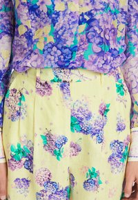 Blumenbluse mit lila und blauen Hortensienmustern; kombiniert mit hellgelben Hosen, die ein ähnliches Blumenmuster und grüne Akzente aufweisen.