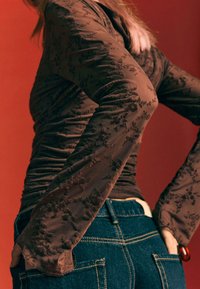 Smeđa majica dugih rukava s cvjetnim vezom i nabranom teksturom, uparena s tamnoplavim trapericama koje imaju stražnje džepove i žute šavove.