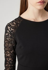 Czarna sukienka z dopasowanym topem i długimi rękawami, wykonana z misternie oprawionej czarnej koronki z wzorami kwiatowymi, kontrastującymi z gładką przednią powierzchnią.