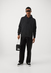 Black hoodie with a front pocket, black bag with a logo, dark denim pants, and black sneakers. Matte sunglasses complete the look.