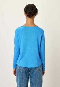 Mėlynos spalvos ilgomis rankovėmis marškiniai, pagaminti iš lengvos medžiagos, su apvaliu iškirpimu. Nugara turi šiek tiek išlenktą apatinę dalį ir tekstūruotą paviršių.