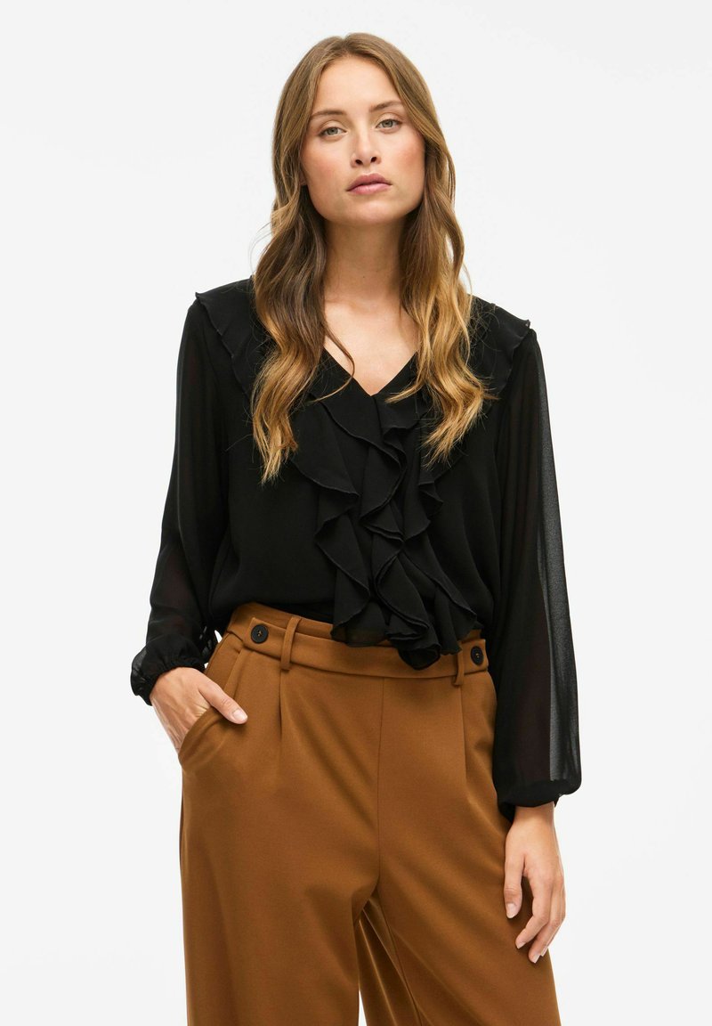 Blusa nera con volant sulla parte anteriore e sui polsini, abbinata a pantaloni marroni a vita alta con due bottoni. Tessuto morbido e design strutturato.