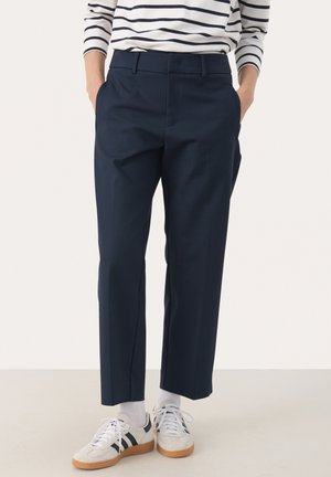 Persoon draagt marineblauwe cropped broek, witte sokken, witte en zwarte sneakers en een wit en marineblauw gestreept shirt met lange mouwen, met handen in de zakken.