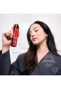 Femme aux longs cheveux foncés tenant une bouteille rouge de L'Oréal Infallible setting mist près de son visage, les yeux fermés, portant un blazer sombre.