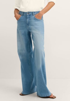 Person trägt eine hellblaue Jeans mit weitem Bein und braune Sandalen mit quadratischer Spitze, Hände in den Taschen, kombiniert mit einem weißen Langarmshirt.