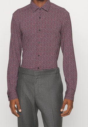 Mies, jolla on päällään istuva viininpunainen kuvioitu napitettava paita ja korkeavyötäröiset harmaat räätälöidyt housut, tasaisen taustan edessä.