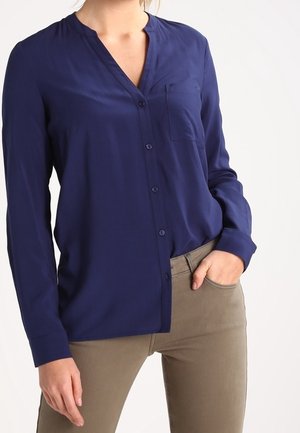 Vrouw draagt een donkerblauw overhemd met lange mouwen, knopen aan de voorkant en een voorzak, en beige broek, hand in de zak.