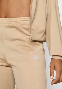 Beige atletisk träningsoverall med ribbat mönster. Har dragkedja och en liten vit broderad logotyp på byxorna.