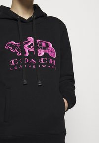 Sudadera negra con bolsillo canguro frontal, que presenta un gráfico brillante en rosa de un dinosaurio y un carro, y detalles con cordones.