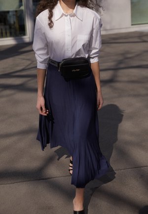 Kobieta spacerująca na zewnątrz w białej koszuli zapinanej na guziki, granatowej plisowanej spódnicy, czarnych sandałkach na obcasie z paskami oraz czarnej torebce na pasku marki Tommy Hilfiger.