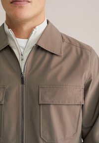 Primo piano di una persona che indossa una giacca color taupe con una grande tasca frontale e una cerniera, sovrapposta a un maglione bianco con cerniera.