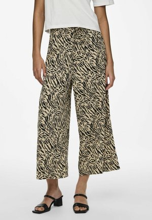 Femme portant un pantalon beige à jambes larges avec un imprimé tigre noir, associé à des sandales à talons noires et un top court blanc.
