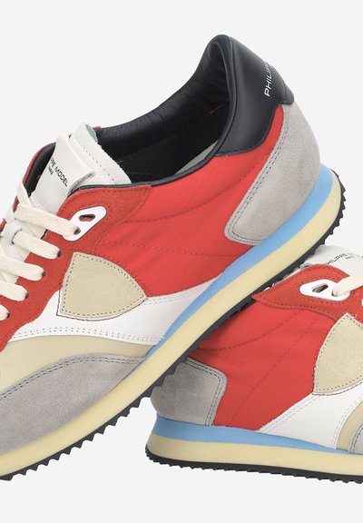 Paire de baskets rouges, blanches, beiges et grises avec des semelles intermédiaires bleues, des semelles extérieures noires, des lacets blancs et des colliers de talon noirs affichant le nom de la marque.