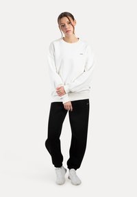 Sudadera blanca de gran tamaño con puños acanalados, combinada con pantalones deportivos negros y zapatillas deportivas blancas. Diseño minimalista, texturas lisas.