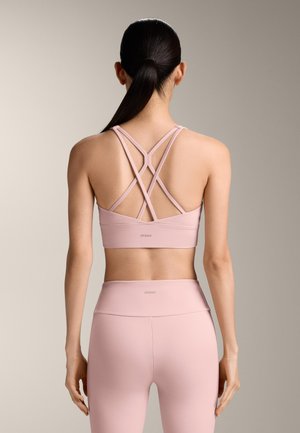 Žena s tamnom kosom u niskom repu, odjevena u ružičasti sportski grudnjak s ukrštenim naramenicama i odgovarajuće visokog struka tajice, prikazana s leđa.