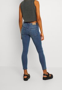 Jeans ajustados en denim azul oscuro con cinco bolsillos, corte de tiro medio y una etiqueta de marca de color claro en la parte trasera. Llevados con sandalias negras.