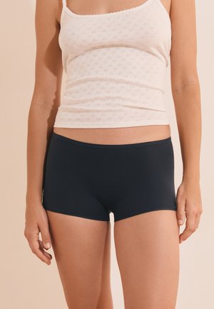 Femme portant un haut blanc sans manches et un short noir ajusté, debout devant un fond beige, les bras détendus le long du corps.