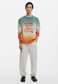 Gebreide trui met een ombre-gradiënt van teal naar oranje, voorzien van geborduurde tekst en zonontwerpen; gecombineerd met gestreepte, losse broek.