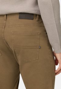 Pantalones de algodón marrón con una textura suave, que cuentan con un bolsillo trasero y una etiqueta de cuero con "BOGGI MILANO" grabada.
