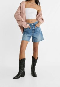 Stivali da cowboy neri, pantaloncini di jeans blu, top bianco corto e una camicia oversize rosa chiaro con tessuto testurizzato e taglio morbido.