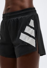 Shorts de sport noirs avec un motif en maille géométrique blanche sur le côté, portés par une personne à peau foncée, montrant la zone abdominale et la partie supérieure de la cuisse.