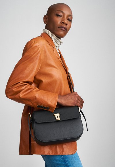 Black leather crossbody bag with a textured finish, gold hardware, and a flap closure, held by a person in an orange leather jacket.