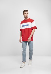 Rood en wit kleurgebroken t-shirt met "STARTER" logo, korte mouwen, gecombineerd met slim-fit lichtblauwe jeans en witte sneakers.