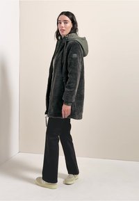 Cappotto in pile nero con cappuccio e superficie testurizzata, con zip frontale e cordino. Indossato sopra pantaloni neri e sneakers chiare.