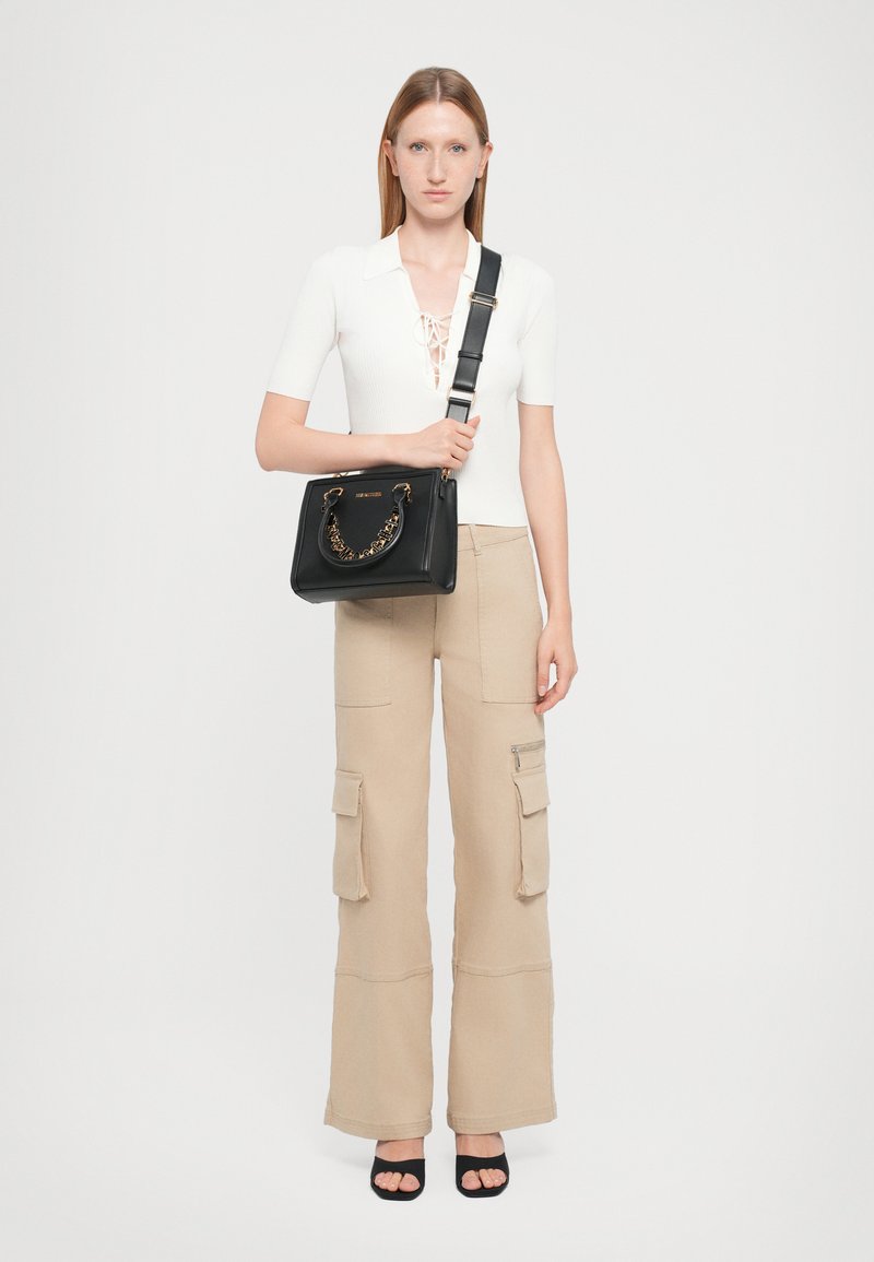Schwarze Lederhandtasche mit Kettenhenkel, quadratischer Form und Reißverschluss. Das Modell trägt ein enges weißes Oberteil und beige Cargo-Hosen. Schwarze Schuhe.