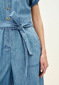 Combinaison en denim bleu clair à manches courtes, fermeture par boutons et ceinture nouée à la taille. Tissu texturé avec poches latérales.