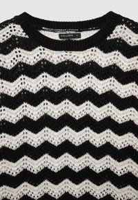Schwarz-weiß gestrickter Pullover mit Zickzackmuster, runder Ausschnitt und zarten Durchbrüchen. Markenetikett sichtbar.