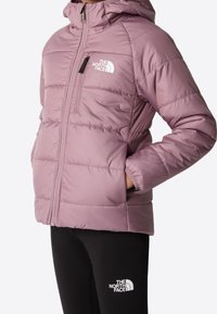 Dziecko w fioletowej kurtce puchowej z kapturem i czarnych spodniach, obie z logo The North Face, z rękami w kieszeniach kurtki.