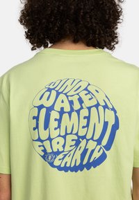Lysegrøn t-shirt lavet af bomuld, med et stort blåt psykedelsk tekstdesign på ryggen med ordene: "Vind, Vand, Jord, Ild, Element."