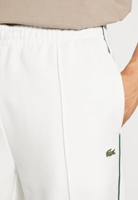 Pantalones de chándal blancos con cintura elástica, bolsillos laterales, rayas de acento verdes y un pequeño logo bordado en el muslo.