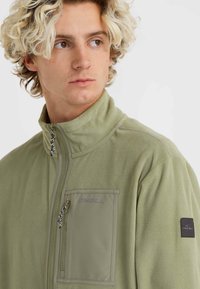 Olive green fleece jacket with high collar, zippered front, and textured fabric. Features a chest pocket and branded accents on the sleeve.