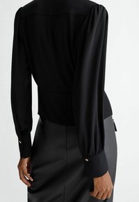 Vista posteriore di una persona che indossa una blusa nera a maniche lunghe con polsini abbottonati e una gonna di pelle nera.