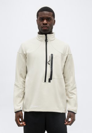 Homme portant un pull léger beige clair avec une poche poitrine zippée verticale noire et un pantalon noir, debout devant un fond blanc uni.