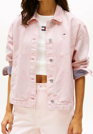 Frau trägt eine hellrosa Jeansjacke mit hochgekrempelten Ärmeln, ein weißes Cropped-Top mit Logo und passende pinke Hose.