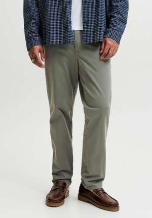 Homme portant un pantalon vert olive, des mocassins en cuir marron et une chemise à carreaux bleue avec les mains tatouées visibles sur les côtés.