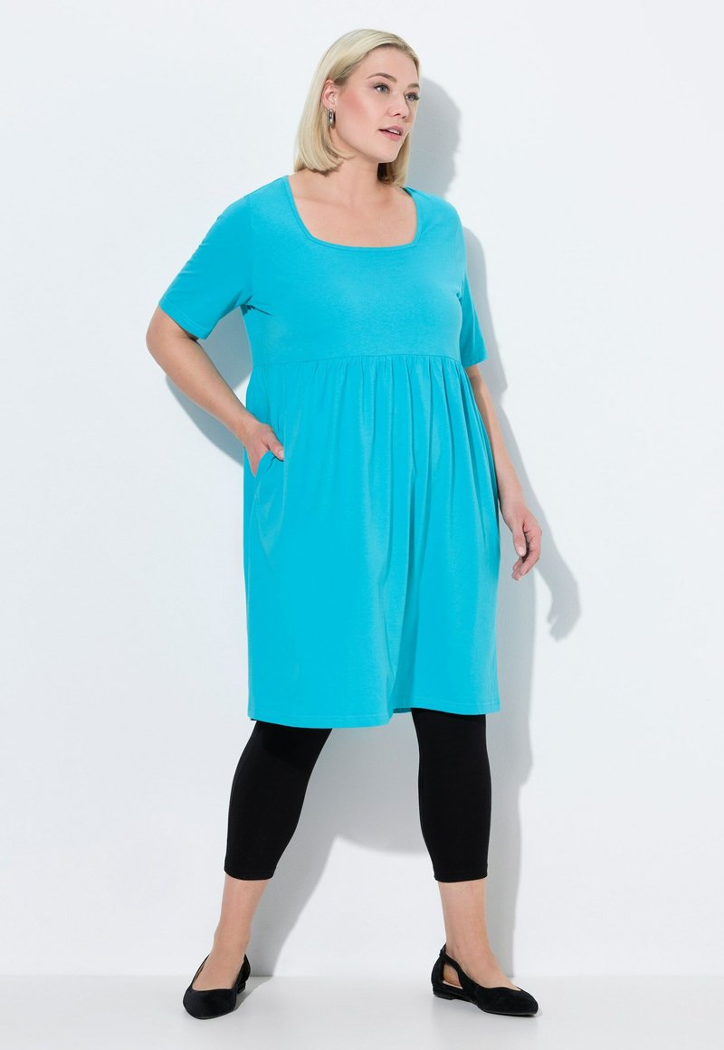 Un vestido turquesa a la altura de la rodilla con mangas cortas y escote cuadrado, que presenta una cintura fruncida y bolsillos laterales, combinado con leggings negros.