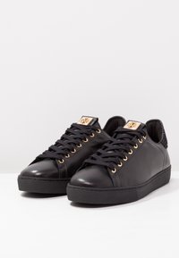 Svarta lädersneakers med guldöglor, svarta snören och en strukturerad bakdel. Slät yta och gummisula. Minimalistisk design.