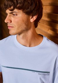T-shirt en coton bleu clair avec un col rond et un graphique horizontal avec du texte en bleu foncé et vert foncé, sur fond en bois.