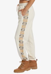 Pantalones de chándal de crema clara con una franja lateral estampada en negro y dorado, que cuentan con una cinturilla y puños elásticos, combinados con botas de senderismo marrones.