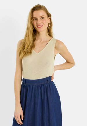 Mujer sonriente con cabello largo y rubio lleva una camiseta de punto sin mangas beige y una falda azul oscuro de talle alto, de pie contra un fondo blanco.