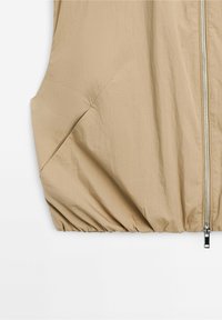 Beige sleeveless vest with front metal zipper, angled side pocket, and gathered elastic hem at the bottom.