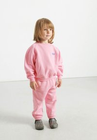 Felpa rosa in cotone con maniche a costine e pantaloni abbinati. Presenta un piccolo logo blu. Abbinata a scarpe slip-on grigie con fori.