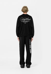 Sudadera negra con texto blanco en la parte posterior, complementada con pantalones negros que presentan acentos gráficos y un detalle de cadena en el lateral.
